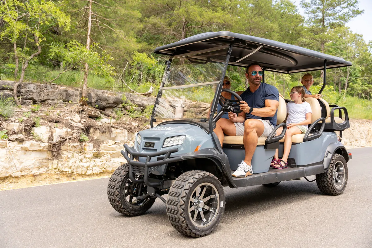 A family rides together in a golf cart along a scenic road surrounded by trees and natural beauty. The group looks cheerful and engaged, clearly enjoying exploration and bonding in the great outdoors.