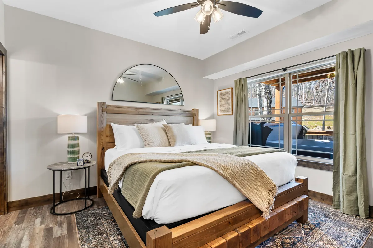 A king-sized bed sits at the center of a tranquil bedroom, topped with plush bedding and decorative pillows. A circular mirror hangs above the bed, large windows with curtains allow soft natural light to filter in, and a bedside lamp adds to the cozy atmosphere.