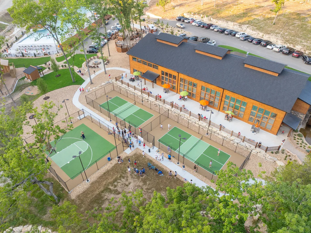 An energetic exterior scene featuring a modern building with expansive sports courts for basketball and tennis. People are actively engaged in games, while others relax in an adjacent seating area. The setting is open and lively, surrounded by greenery.