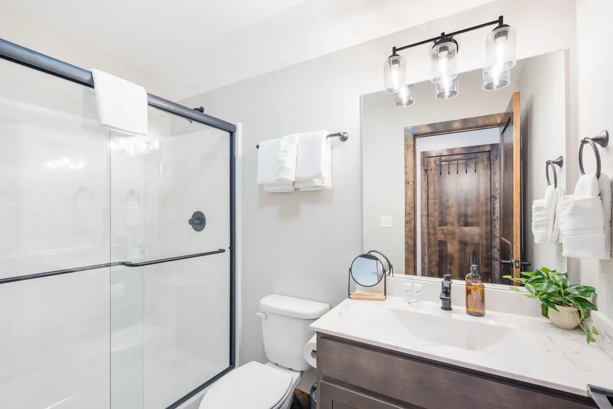 A modern ensuite bathroom with a glass shower enclosure, a spacious vanity with sink, a decorative mirror, and stylish light fixtures. The space is bright, fresh, and inviting.