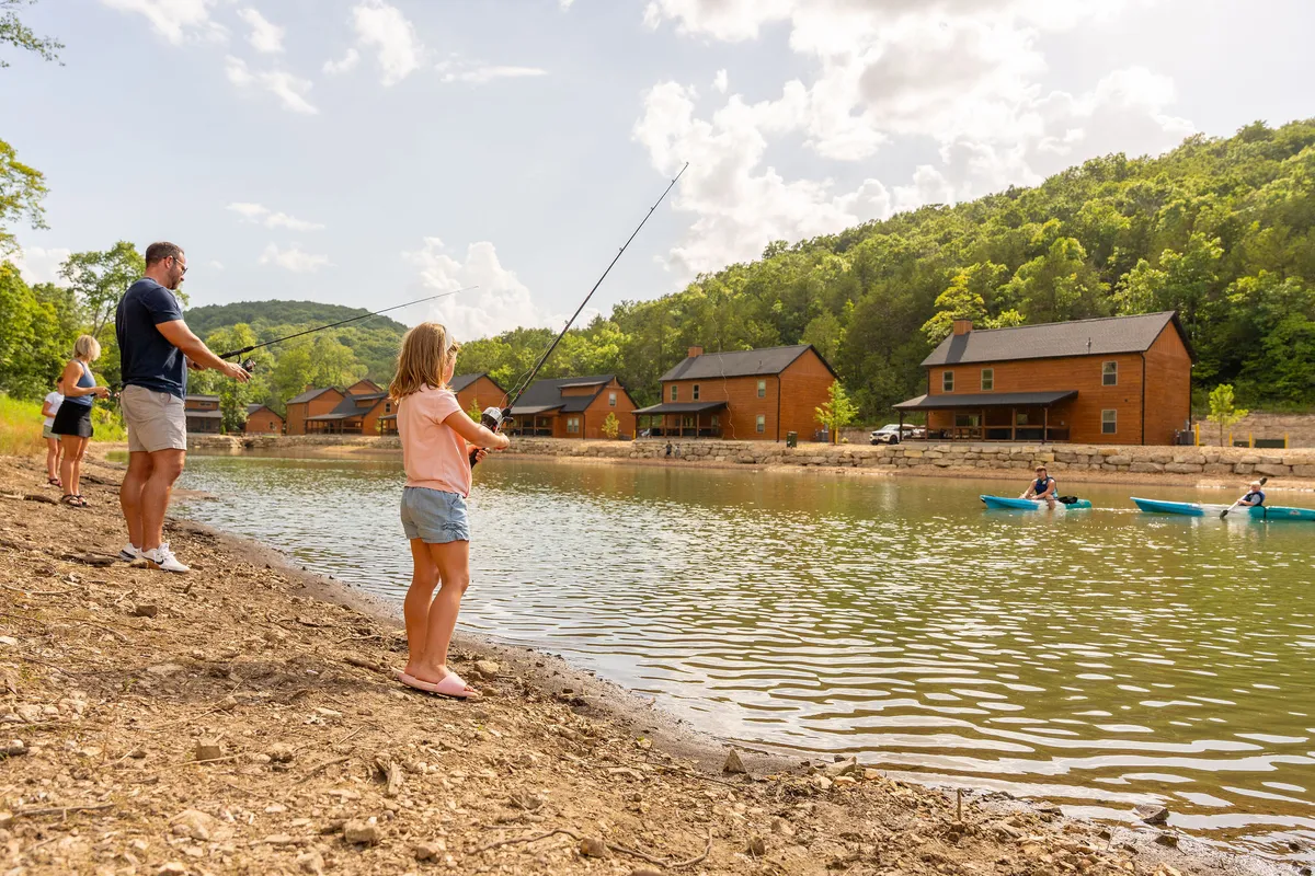 A joyful family scene at the lake, with a child fishing alongside other family members, cabins visible nearby, and kayakers gliding across the water's surface. The sun is shining, and the overall atmosphere is lively, with people actively engaging in a variety of lakeside activities.