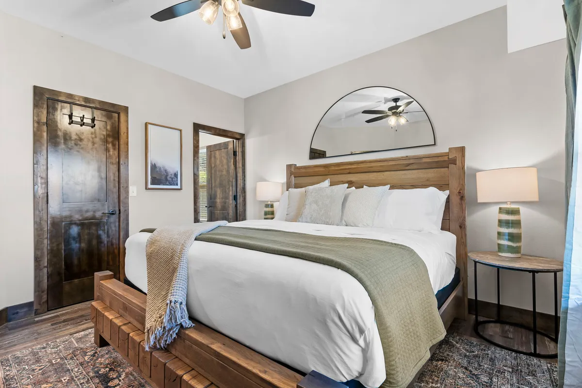 A comfortable bedroom featuring a wooden bed frame, textured throw blanket, and nightstands with lamps. Decorative elements like a mirror above the bed and a warm, neutral color palette create an inviting and peaceful space.