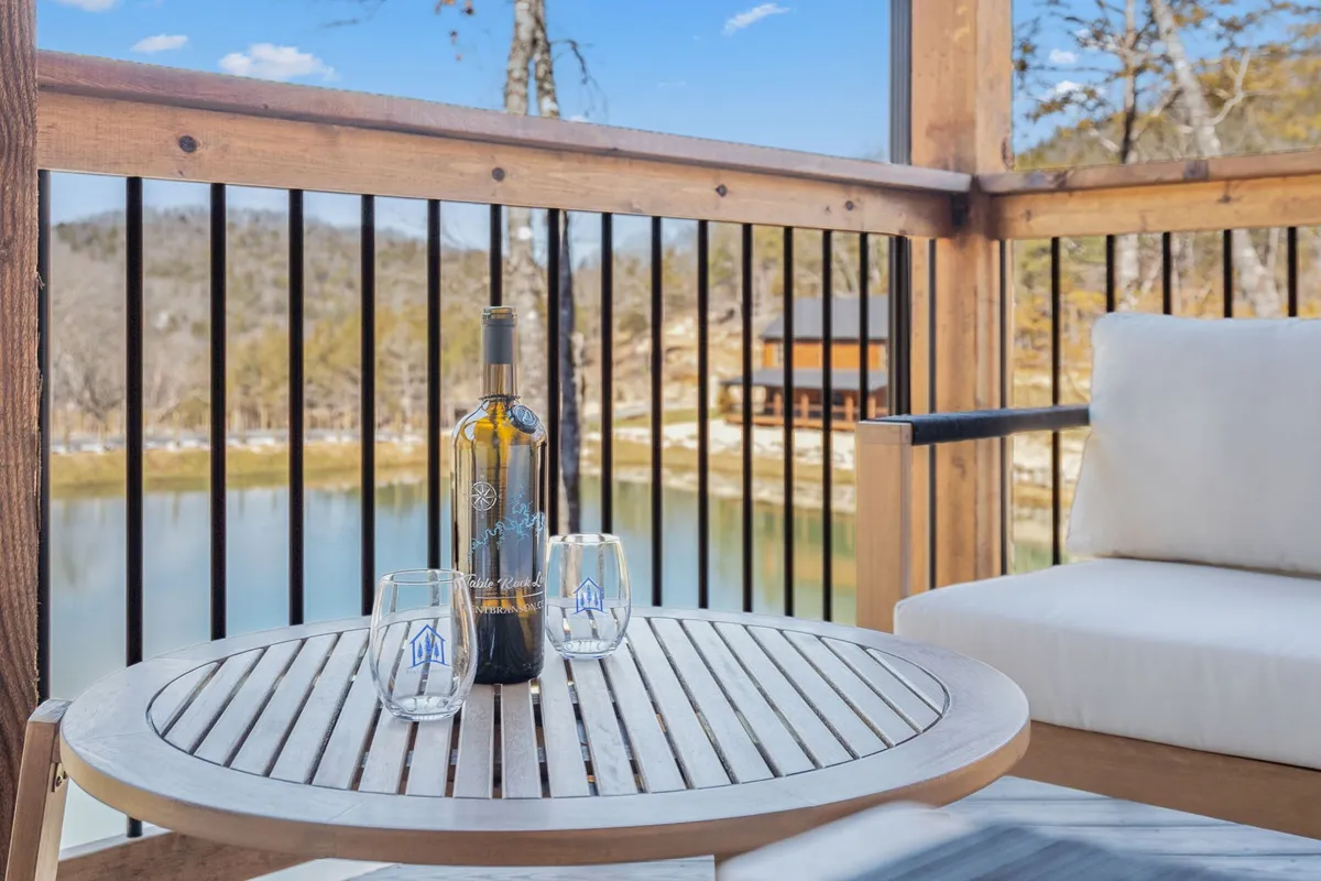 A wooden outdoor table set with a bottle of wine and two glasses, positioned to enjoy a breathtaking scenic view over the water. The mood is intimate and inviting, perfect for savoring a drink as the sun sets.