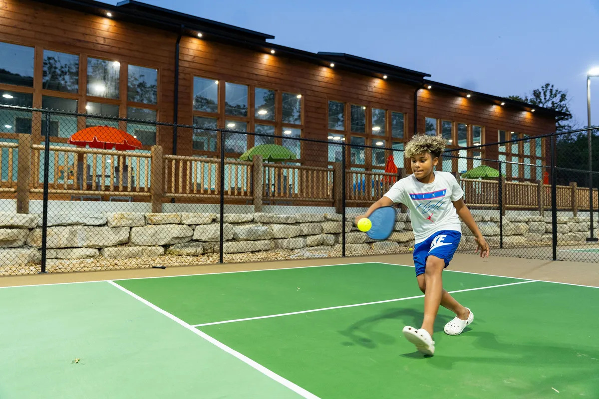 A child plays pickleball on a tennis court at sunset, with a pool area visible in the background. The scene is energetic and joyful, highlighting family-oriented recreation and the new sports amenities at Tall Timbers Camp.