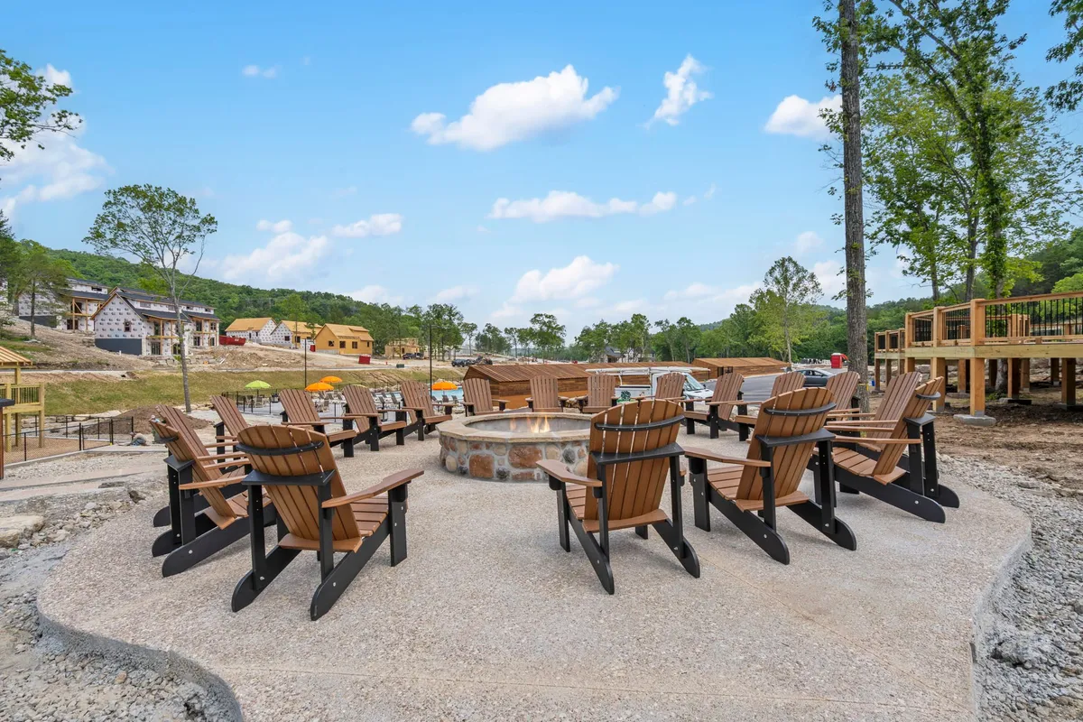 A cozy fire pit is encircled by adirondack chairs, set amidst trees with a hint of construction in the background. The space feels both rustic and communal, inviting guests to gather, share stories, and enjoy nature, even mentioning the ongoing development that new visitors might encounter.