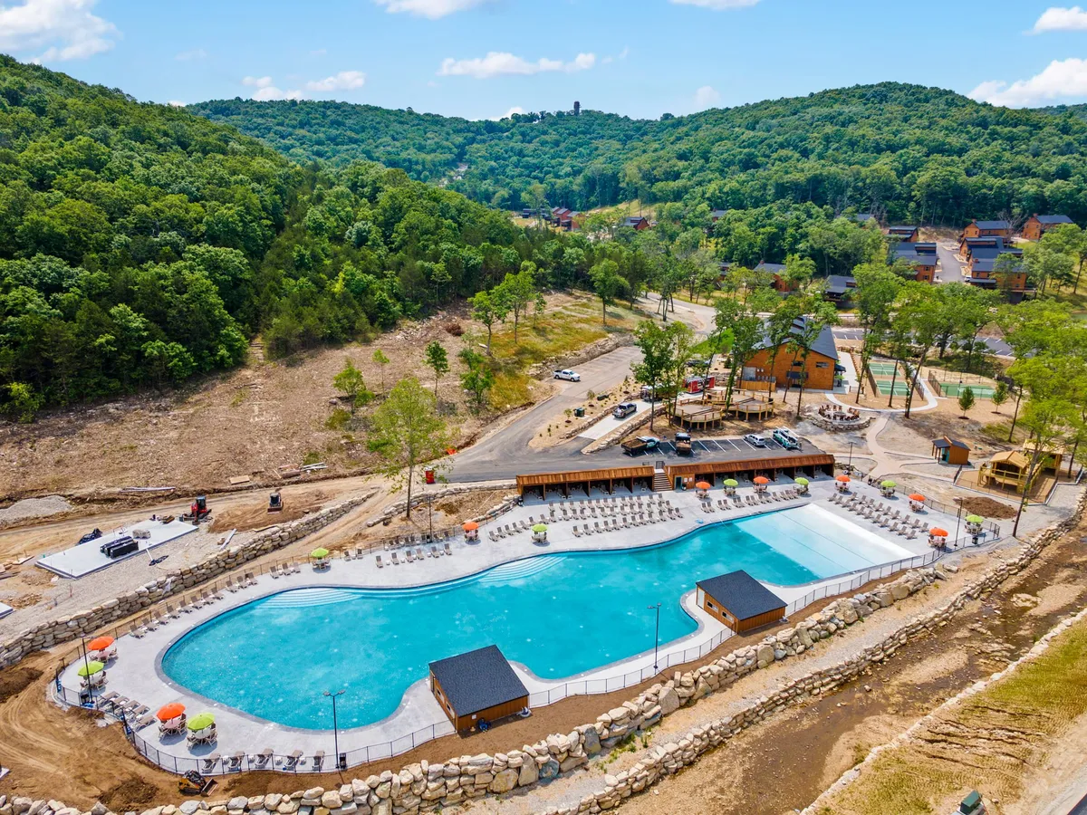 A large, cheerful outdoor swimming pool area with lounge chairs, umbrellas, a wooden deck, and nearby cabins nestled against green hills. The setting feels vibrant and communal, perfect for families and groups to gather, swim, and relax throughout the day.