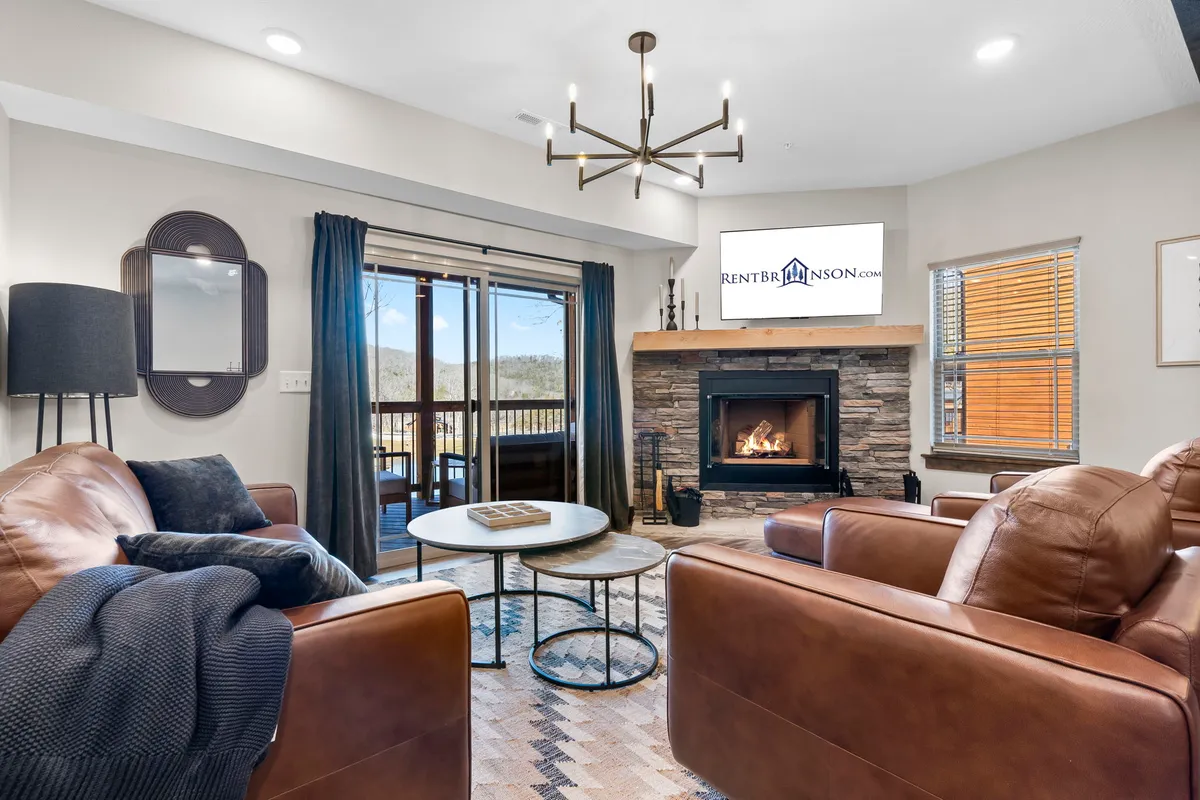 A spacious and comfortable living room featuring leather sofas, a stone fireplace, a coffee table, and large windows overlooking scenic outdoor views. The arrangement is ideal for socializing, relaxing by the fire, or enjoying the surrounding beauty through the windows.