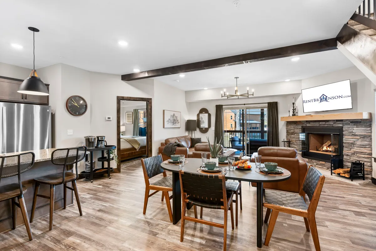 The image showcases a warmly lit interior featuring a rustic dining table set for a meal, a cozy stone fireplace, a modern kitchen island, and a plush seating area. The space exudes comfort and is arranged to invite gatherings, relaxation, and time together.