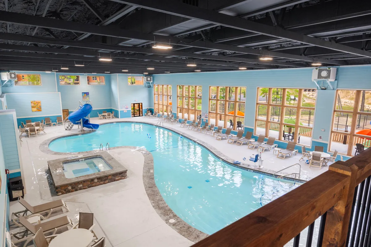 A bright indoor swimming pool area featuring a water slide, hot tub, lounge chairs, and large windows that offer views of the outdoors. The space feels fun, lively, and perfect for all-weather enjoyment.