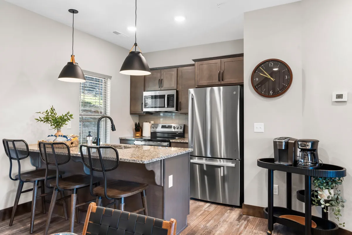 This image features a modern kitchen with a granite countertop, sleek appliances, and bar stools that invite guests to sit and socialize. A decorative clock adds a homey touch, while the overall layout showcases both style and functionality.