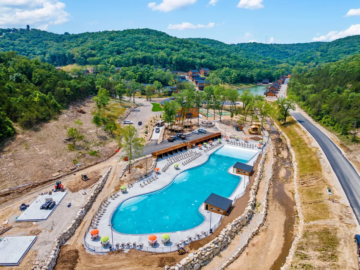 A large, inviting outdoor swimming pool is surrounded by green hills, lounge chairs, and a wooden building with a deck. The scene is set at sunset, capturing a vibrant yet relaxed mood perfect for group activities and social gatherings.